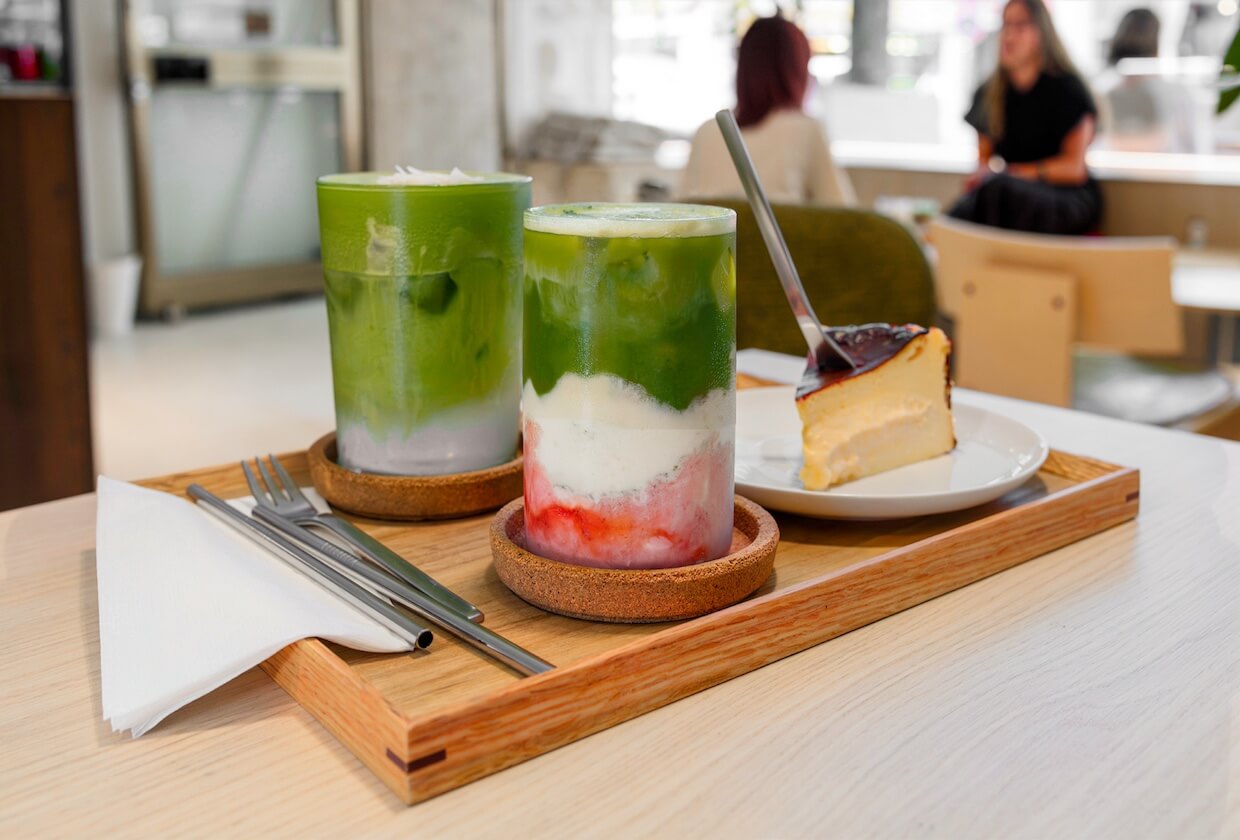 Zwei Matcha-Drinks, einer davon mit Coconut und einer mit Erdbeere, und ein Stück baskischer Käsekuchen auf einem Holztablett