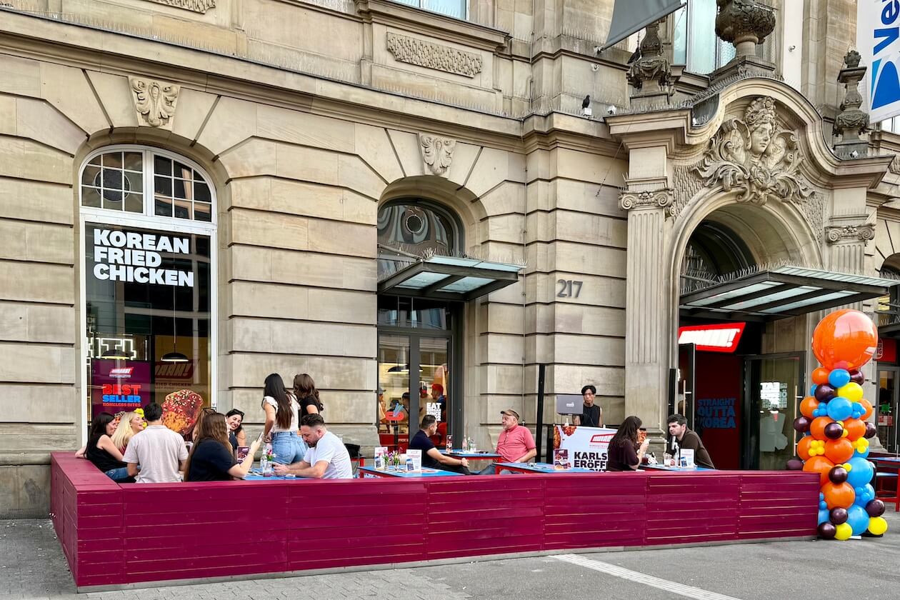 Außenansicht von Mmaah! Karlsruhe in der historischen Postgalerie, Kaiserstraße 217, mit roter Terrasse, Gästen und Ballonsäule zur Eröffnung