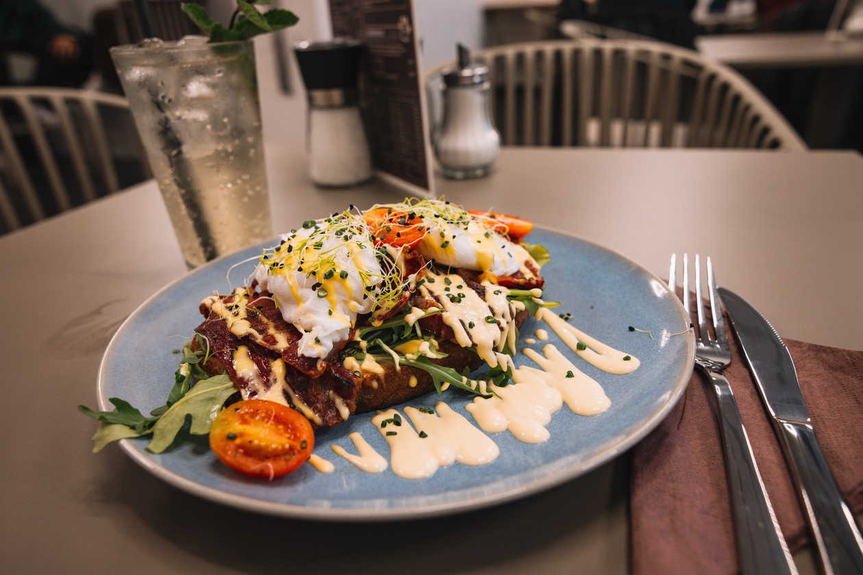 Bacon Eggs Benedict Deluxe auf blauem Teller: geröstetes Sauerteigbrot mit Bacon, pochierten Eiern, Sauce hollandaise, Rucola, Kresse und Kirschtomaten