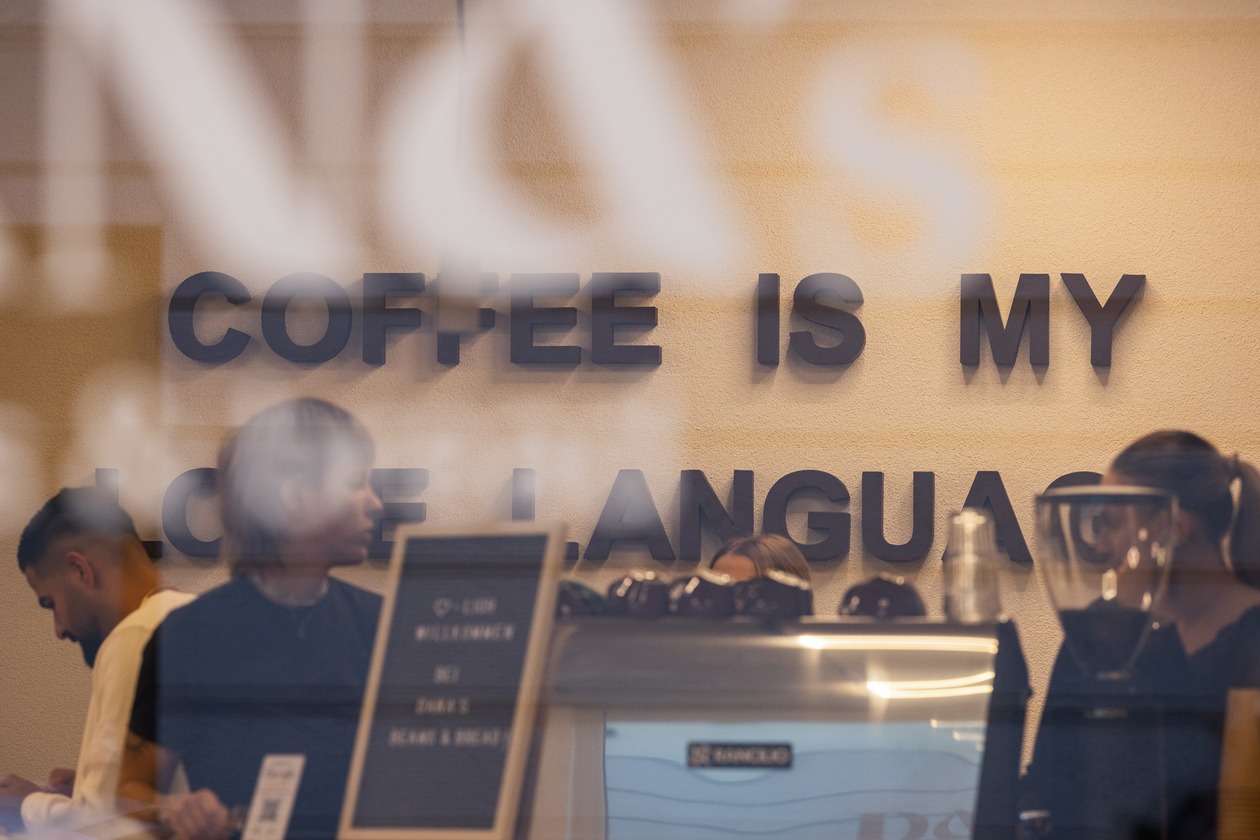 Schriftzug „Coffee is my love language“ an der Wand hinter der Theke, durch das Schaufenster fotografiert, davor Gäste und Mitarbeitende