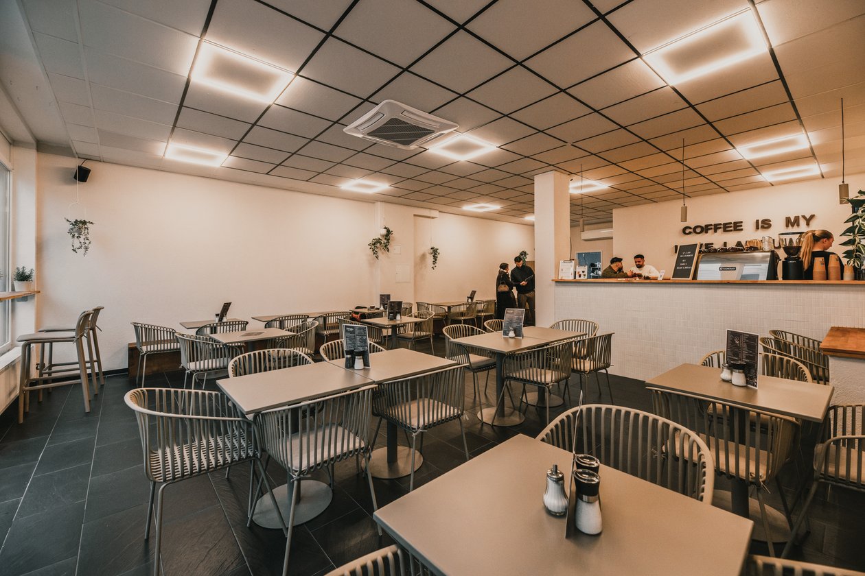 Heller Gastraum von Dana's Beans & Bread mit grauen Tischen, geflochtenen Stühlen, Lichtpaneelen an der Decke und der Theke im Hintergrund