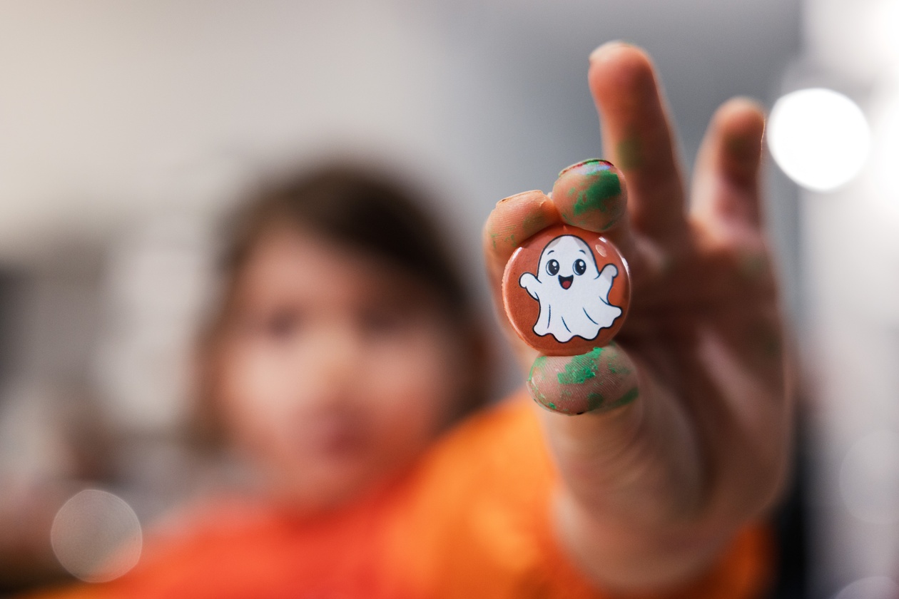 Kind hält einen orangen Button mit Geistermotiv in die Kamera, Finger voller grüner Farbe
