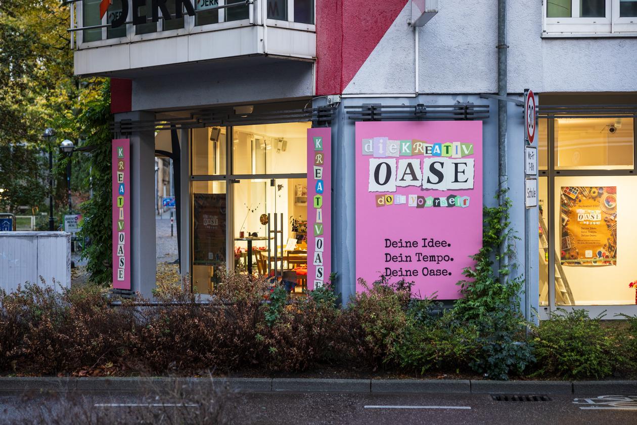 Pinkes Schild der Kreativoase Karlsruhe mit dem Slogan „Deine Idee. Dein Tempo. Deine Oase.“ vor beleuchteten Schaufenstern