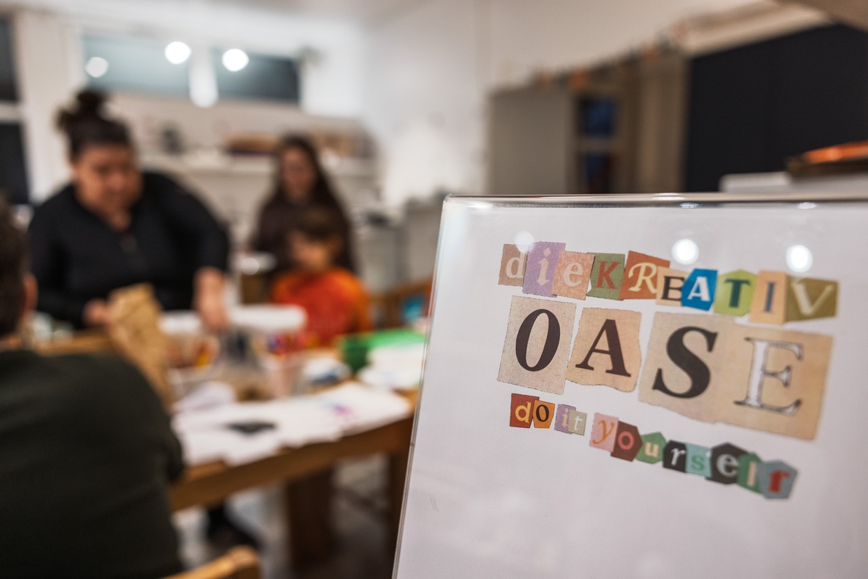 Logo der Kreativoase im Collage-Stil, im unscharfen Hintergrund basteln Gäste am Tisch